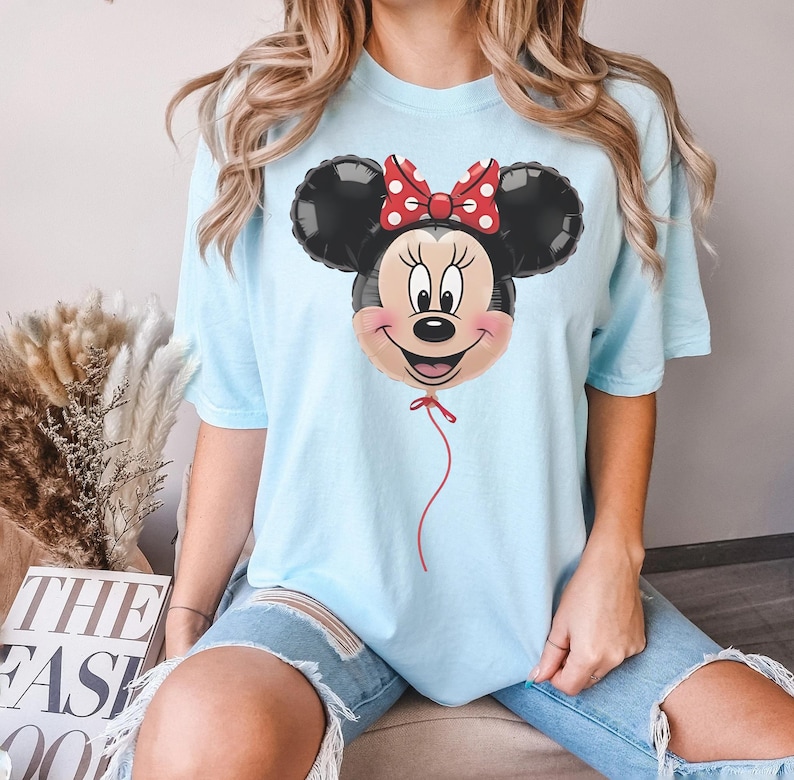May include: Light blue t-shirt featuring a Minnie Mouse balloon graphic. The balloon has black ears, a red bow with white polka dots, and a smiling face. A red string hangs from the balloon. The shirt is a light blue color.