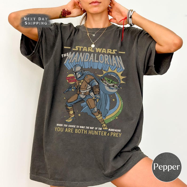 May include: Dark gray t-shirt featuring a Star Wars The Mandalorian graphic. The design includes the Mandalorian, Grogu, and text that reads "When you choose to walk the way of the Mandalore, you are both hunter & prey."
