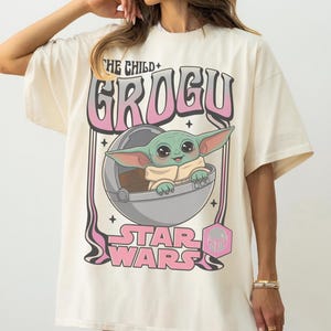 Puede incluir: Una camiseta color hueso con un gr&aacute;fico de Grogu de Star Wars dentro de una c&aacute;psula flotante. El texto dice THE CHILD GROGU y STAR WARS en letras rosas y negras. El dise&ntilde;o incluye peque&ntilde;as estrellas negras y un icono hexagonal rosa.