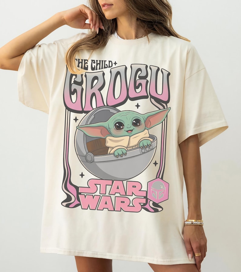 Puede incluir: Una camiseta color hueso con un gr&aacute;fico de Grogu de Star Wars dentro de una c&aacute;psula flotante. El texto dice THE CHILD GROGU y STAR WARS en letras rosas y negras. El dise&ntilde;o incluye peque&ntilde;as estrellas negras y un icono hexagonal rosa.
