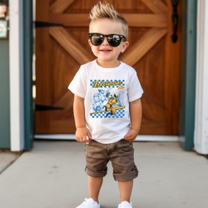 May include: A young child wearing a white t-shirt with a Wolverine graphic and the word "Wolverine" in blue and yellow. The child is also wearing brown shorts and sunglasses.