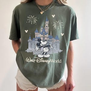 May include: Olive green t-shirt featuring a graphic of Mickey Mouse in front of Cinderella's castle at Walt Disney World. The design includes fireworks and the text "Walt Disney World" in white. The shirt has short sleeves.