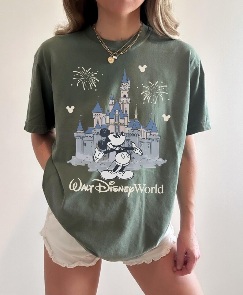 May include: Olive green t-shirt featuring a graphic of Mickey Mouse in front of Cinderella's castle at Walt Disney World. The design includes fireworks and the text "Walt Disney World" in white. The shirt has short sleeves.
