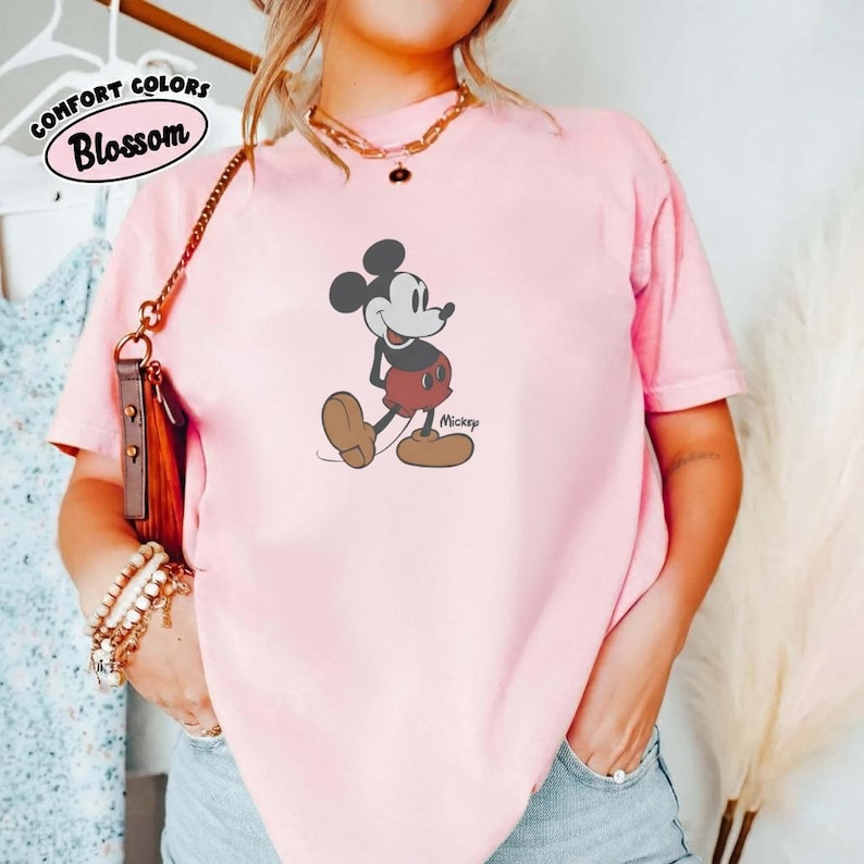 Op de afbeelding: Een lichtroze t-shirt met een Mickey Mouse-afbeelding. Het stripfiguur wordt getoond in zijn klassieke pose, met een rode korte broek en bruine schoenen. Het shirt heeft ook de tekst "Comfort Colors Blossom" in een cirkelvormig ontwerp.