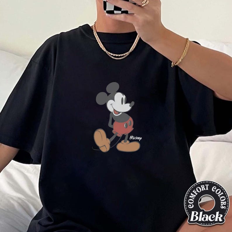 Op de afbeelding: Zwart T-shirt met een cartoonillustratie van Mickey Mouse. Mickey wordt afgebeeld in een loophouding, met een rode broek en gele schoenen. Het shirt heeft een Comfort Colors-label rechtsonder.