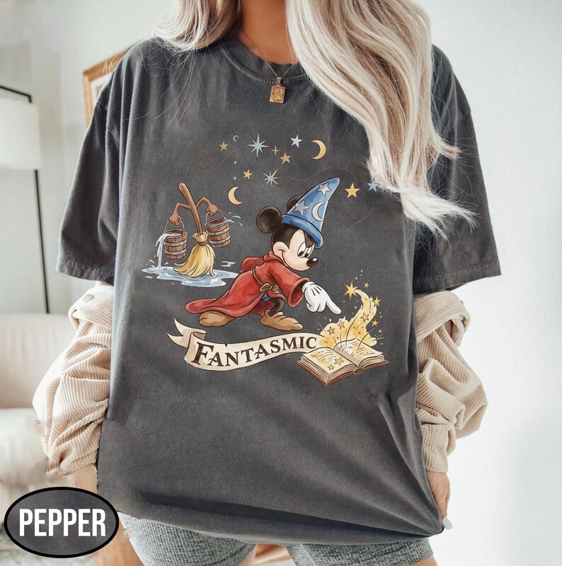 May include: Dark gray t-shirt featuring a cartoon Mickey Mouse dressed as a sorcerer, with a red robe and blue hat. The design includes a broom, stars, moons, and the word "Fantasmic" on a banner.