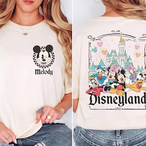 May include: Ivory t-shirt with a Mickey Mouse graphic and the name "Melody". The back features a Disneyland design with cartoon characters and the text "Disneyland 1955". The shirt is a cream colour.