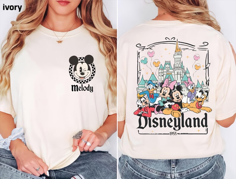 May include: Ivory t-shirt with a Mickey Mouse graphic and the name "Melody". The back features a Disneyland design with cartoon characters and the text "Disneyland 1955". The shirt is a cream colour.
