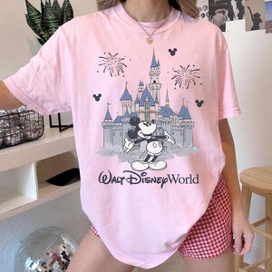 May include: Pink t-shirt featuring a graphic of Mickey Mouse in front of a castle with fireworks and the words "Walt Disney World". The shirt has short sleeves and a crew neck.