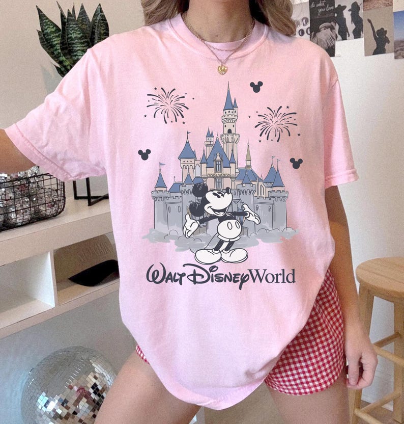 May include: Pink t-shirt featuring a graphic of Mickey Mouse in front of a castle with fireworks and the words "Walt Disney World". The shirt has short sleeves and a crew neck.