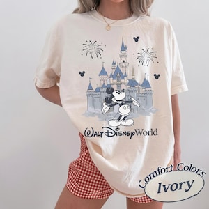 May include: Ivory-colored t-shirt featuring a graphic of Mickey Mouse in front of a blue castle with fireworks. The text "Walt Disney World" is below the graphic. The shirt is labeled "Comfort Colors Ivory".