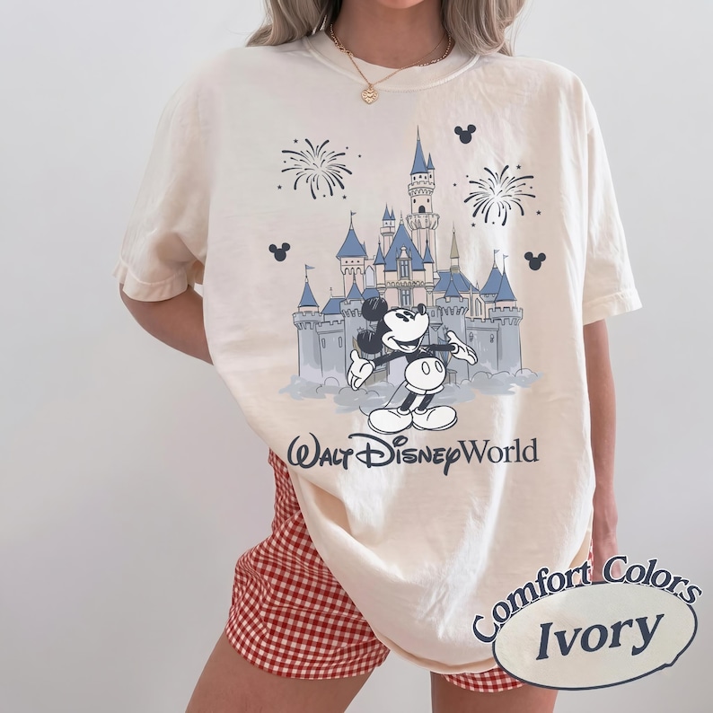 May include: Ivory-colored t-shirt featuring a graphic of Mickey Mouse in front of a blue castle with fireworks. The text "Walt Disney World" is below the graphic. The shirt is labeled "Comfort Colors Ivory".