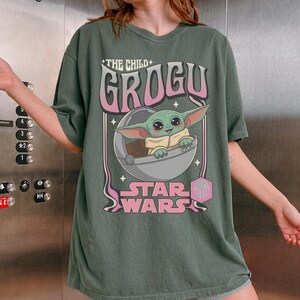 Puede incluir: Una camiseta de manga corta en color verde oliva oscuro con un estampado gr&aacute;fico del personaje Grogu de Star Wars dentro de una c&aacute;psula flotante. El texto dice The Child, Grogu y Star Wars en una fuente retro con detalles en rosa y blanco.