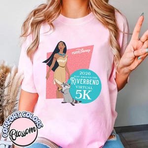 May include: Pink t-shirt featuring a graphic of Pocahontas, a raccoon, and text that reads "Run Disney" and "2026 Just Around the Riverbend Virtual 5K." The shirt also has the "Comfort Colors Blossom" logo.