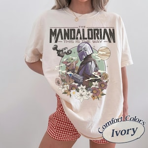 May include: Ivory-colored t-shirt featuring a graphic of The Mandalorian and Grogu, surrounded by flowers and planets. The text "THE MANDALORIAN - THIS IS THE WAY" is printed above the characters. The shirt is a Comfort Colors brand.