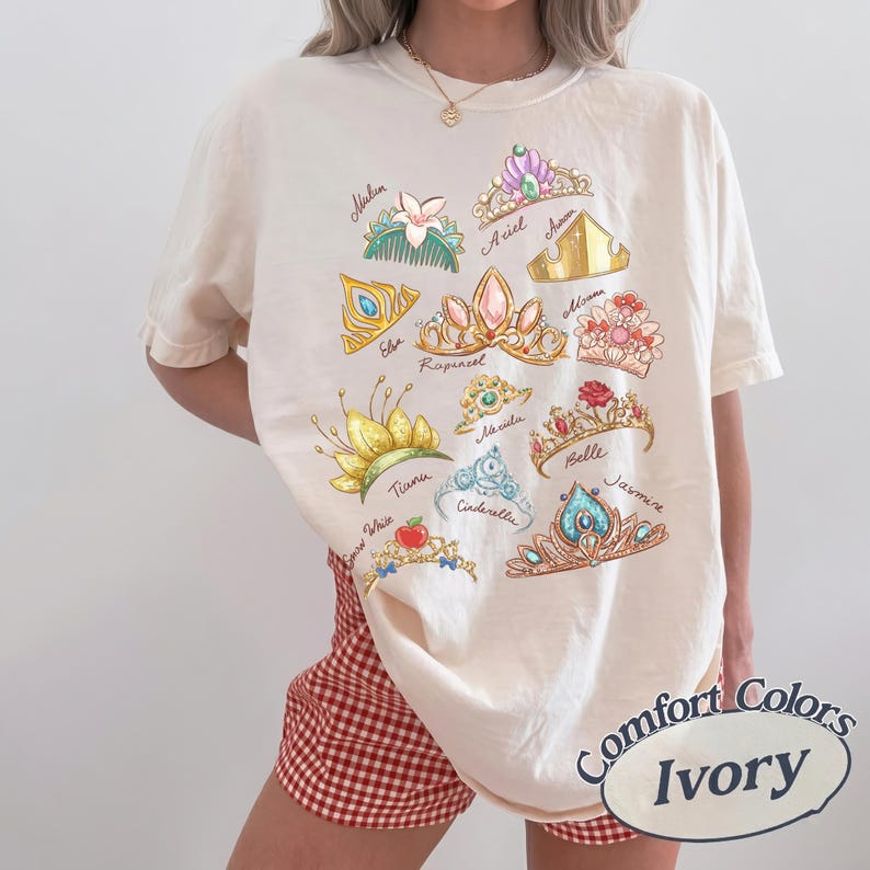 May include: Ivory-colored t-shirt featuring colorful illustrations of princess tiaras with names like Ariel, Belle, and Jasmine. The shirt has a Comfort Colors label. The model is wearing red and white checkered shorts.