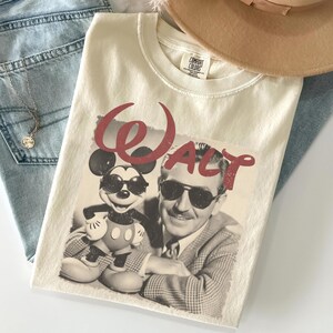 Pu&ograve; includere: T-shirt color crema con grafica di Topolino e Walt Disney, con la scritta "Walt" in rosso. La maglietta &egrave; piegata sopra jeans blu, con una collana e un cappello beige visibili.