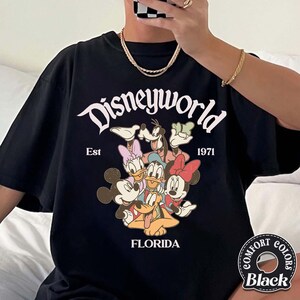 May include: Black t-shirt featuring a Disney World graphic with Mickey Mouse, Minnie Mouse, Donald Duck, Daisy Duck, Goofy, and Pluto. The text "Disneyworld Florida Est 1971" is displayed in a vintage style. The shirt is a Comfort Colors brand.