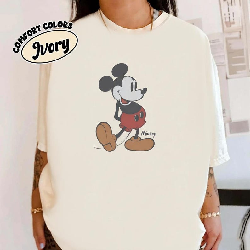 Op de afbeelding: Ivoor T-shirt met een Mickey Mouse-afbeelding. Het stripfiguur is afgebeeld in een rode broek, bruine schoenen en zwarte oren. Op het shirt staan ook de woorden "Comfort Colors" en "Ivory".