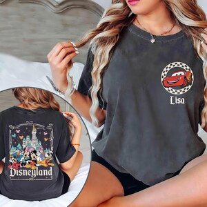 May include: Dark grey t-shirt with a red cartoon race car in a chequered circle, and the name "Lisa" below. The back of the shirt features a Disneyland graphic with cartoon characters and the text "Disneyland 1955".