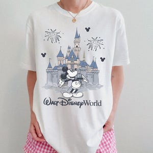 May include: White t-shirt featuring a graphic of Mickey Mouse in front of Cinderella's castle at Walt Disney World. The design includes fireworks and the text "Walt Disney World" in a script font. The shirt is a classic crew neck.
