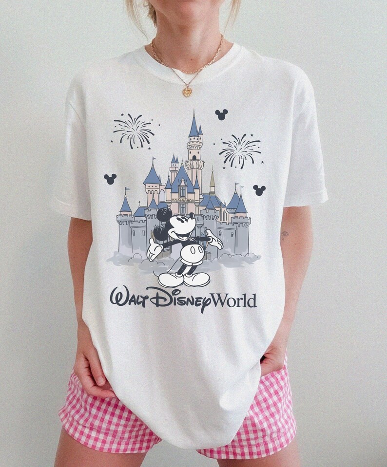 May include: White t-shirt featuring a graphic of Mickey Mouse in front of Cinderella's castle at Walt Disney World. The design includes fireworks and the text "Walt Disney World" in a script font. The shirt is a classic crew neck.