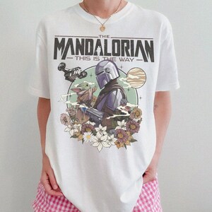 May include: White t-shirt featuring a graphic of The Mandalorian and Grogu, with the text "THE MANDALORIAN THIS IS THE WAY." The design includes a space scene with flowers. The shirt is a crew neck.