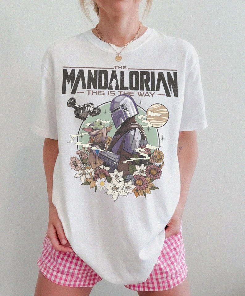 May include: White t-shirt featuring a graphic of The Mandalorian and Grogu, with the text "THE MANDALORIAN THIS IS THE WAY." The design includes a space scene with flowers. The shirt is a crew neck.