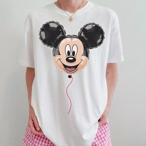 May include: White t-shirt featuring a Mickey Mouse balloon graphic. The balloon has black ears, a tan face, and a red string. The shirt is a casual style, suitable for everyday wear.