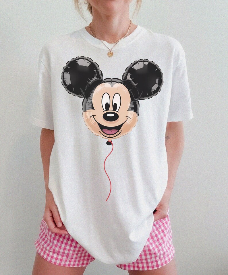 May include: White t-shirt featuring a Mickey Mouse balloon graphic. The balloon has black ears, a tan face, and a red string. The shirt is a casual style, suitable for everyday wear.