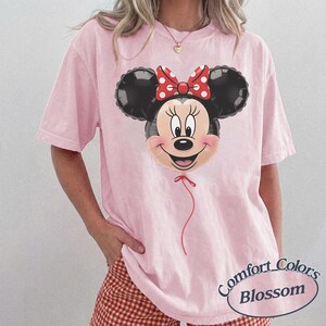 May include: A light pink Comfort Colors t-shirt featuring a Minnie Mouse balloon graphic. The balloon has black ears, a smiling face, and a red bow with white polka dots. The shirt has short sleeves and a relaxed fit.