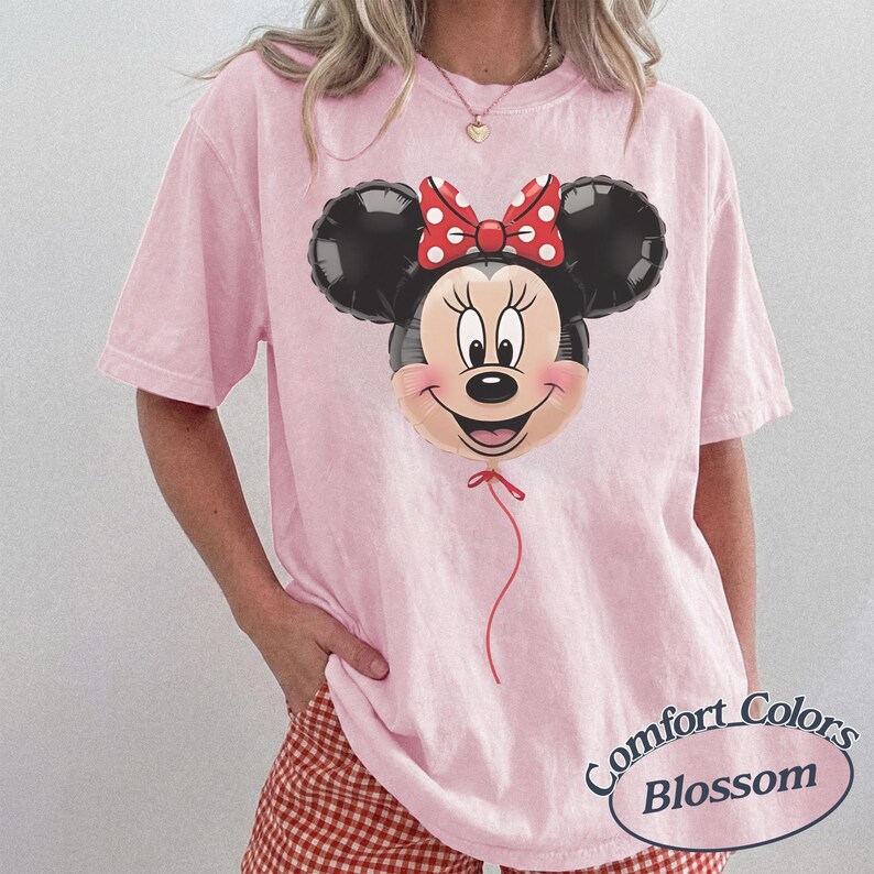 May include: A light pink Comfort Colors t-shirt featuring a Minnie Mouse balloon graphic. The balloon has black ears, a smiling face, and a red bow with white polka dots. The shirt has short sleeves and a relaxed fit.
