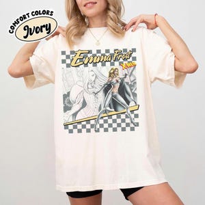 May include: Ivory-colored t-shirt featuring a graphic of Emma Frost from X-Men. The design includes a checkered background, the character's name, and the X-Men logo. The shirt has a relaxed fit and the words "Comfort Colors" are printed on the top left.