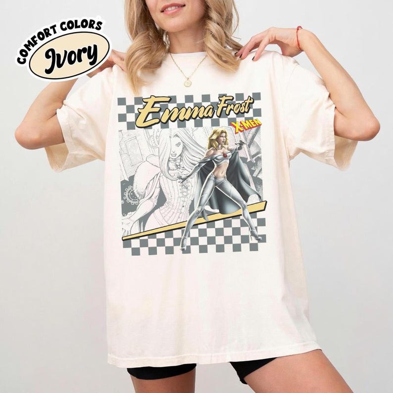 May include: Ivory-colored t-shirt featuring a graphic of Emma Frost from X-Men. The design includes a checkered background, the character's name, and the X-Men logo. The shirt has a relaxed fit and the words "Comfort Colors" are printed on the top left.