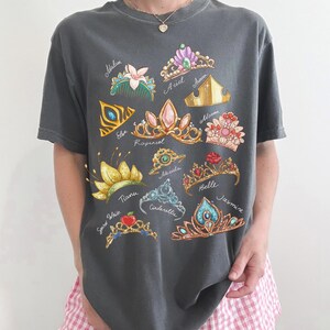 May include: Dark gray t-shirt featuring colorful illustrations of various princess tiaras, each labeled with a name. The tiaras are adorned with gems and floral designs. The shirt is a casual, comfortable style.