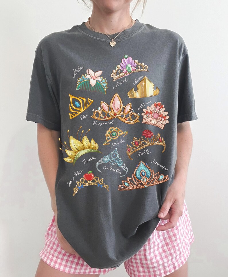May include: Dark gray t-shirt featuring colorful illustrations of various princess tiaras, each labeled with a name. The tiaras are adorned with gems and floral designs. The shirt is a casual, comfortable style.