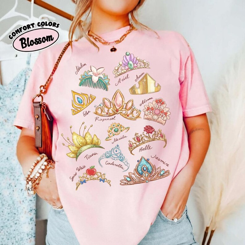 May include: Pink Comfort Colors Blossom t-shirt featuring illustrations of various princess crowns, including Ariel, Belle, and Jasmine. The shirt has a crew neck and short sleeves. The text "COMFORT COLORS" and "Blossom" are printed on the top left.
