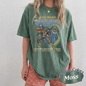 May include: Green "Comfort Colors" t-shirt featuring a graphic of The Mandalorian and Grogu. The design includes the text "STAR WARS THE MANDALORIAN" and "YOU ARE BOTH HUNTER & PREY". The shirt is a moss green color.