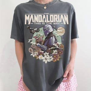 May include: Dark gray t-shirt featuring a graphic of The Mandalorian and Grogu surrounded by flowers, with the text "THE MANDALORIAN - THIS IS THE WAY." The design includes a spaceship and a planet. The shirt is a comfortable, casual style.