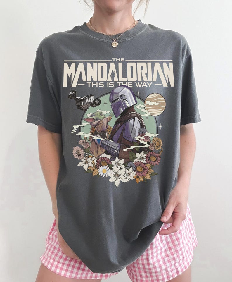 May include: Dark gray t-shirt featuring a graphic of The Mandalorian and Grogu surrounded by flowers, with the text "THE MANDALORIAN - THIS IS THE WAY." The design includes a spaceship and a planet. The shirt is a comfortable, casual style.