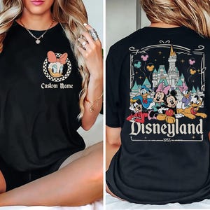 May include: Black t-shirt with Daisy Duck in a pink bow within a chequered circle, and the text "Custom Name". The back shows a Disneyland graphic with Mickey, Minnie, Donald, and Daisy Duck, and the text "Disneyland 1955".