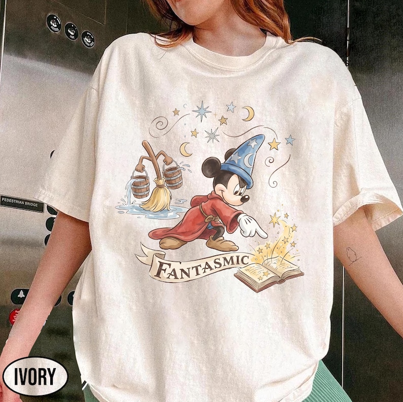 May include: Ivory-colored t-shirt featuring a whimsical illustration of Mickey Mouse dressed as a sorcerer. Mickey is wearing a blue wizard hat and red robe, with the word "Fantasmic" on a banner below. The design includes stars, moons, and a broom.