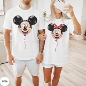 May include: White t-shirts featuring Mickey and Minnie Mouse balloon designs. The Mickey Mouse balloon has a black head and ears, a smiling face, and a red string. The Minnie Mouse balloon has a red bow and a red string. The word "White" is in a white circle in the bottom left corner.