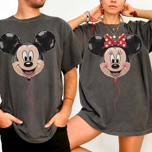 May include: Two charcoal gray t-shirts featuring Mickey and Minnie Mouse balloon designs. Mickey has a smiling face with black ears and a red string. Minnie has a red bow with white polka dots and a red string.