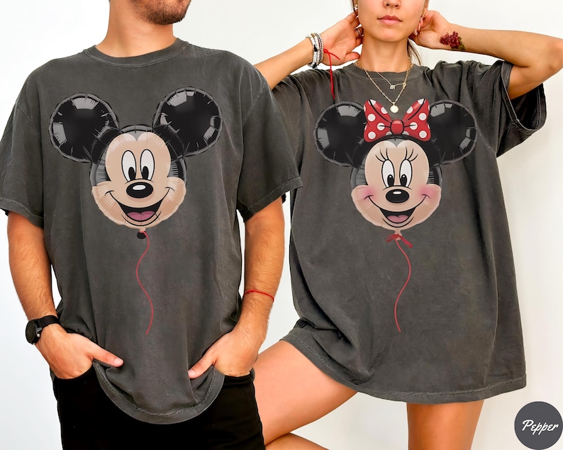 May include: Two charcoal gray t-shirts featuring Mickey and Minnie Mouse balloon designs. Mickey has a smiling face with black ears and a red string. Minnie has a red bow with white polka dots and a red string.
