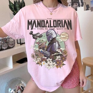 May include: A light pink t-shirt featuring a graphic of The Mandalorian and Grogu, with the text "THE MANDALORIAN THIS IS THE WAY". The design includes a spaceship, flowers, and a planet.