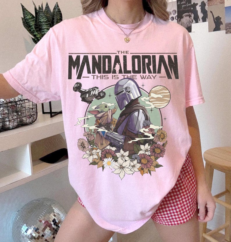 May include: A light pink t-shirt featuring a graphic of The Mandalorian and Grogu, with the text "THE MANDALORIAN THIS IS THE WAY". The design includes a spaceship, flowers, and a planet.
