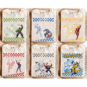 May include: Six folded white t-shirts featuring Marvel comic book characters. Each shirt has a different design, including Hulk, Captain America, Iron Man, Spider-Man, Thor, and Wolverine. Each design includes the character's name and a year.