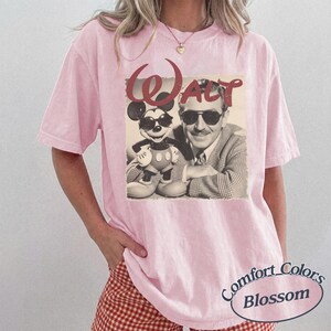 Pu&ograve; includere: T-shirt rosa chiaro con un'immagine vintage in bianco e nero di Walt Disney e Topolino con occhiali da sole. La parola "Walt" &egrave; stampata in rosso sopra l'immagine. La maglietta ha un'etichetta Comfort Colors Blossom.