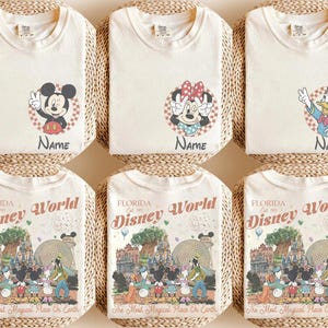 Op de afbeelding: Zes cr&egrave;mekleurige t-shirts met Disney-personage-afbeeldingen. De bovenste rij toont Mickey Mouse, Minnie Mouse en Donald Duck. De onderste rij toont shirts met een Florida Disney World-ontwerp. Elk shirt wordt op een geweven mat gepresenteerd.